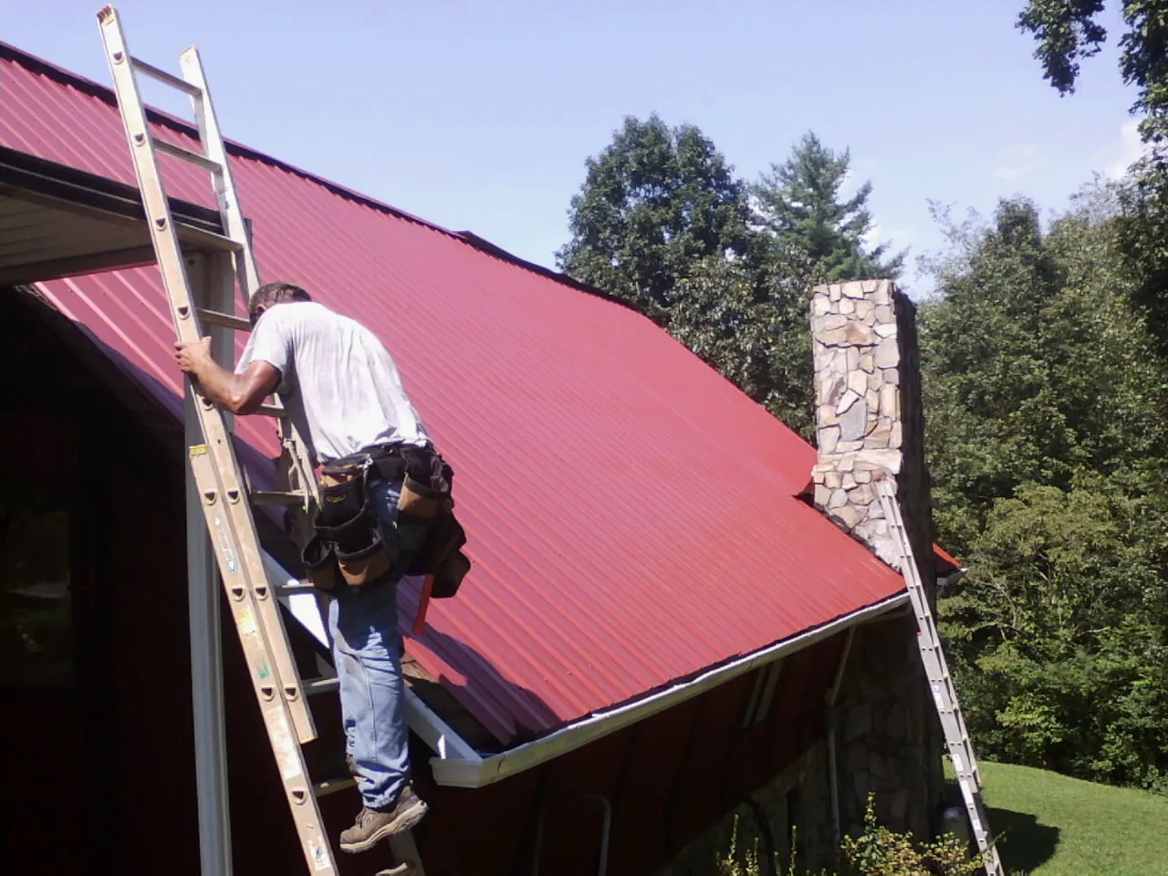 Roofer on a ladder beside a red metal roof and stone chimney — Soffit & Fascia Repair by Roofing Brothers in Sloan